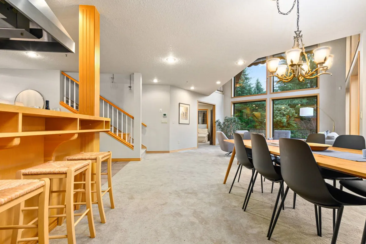 A bright, inviting dining area featuring a large dining table, a bar area, and wide windows that fill the space with natural light. The chandelier overhead and the carefully arranged furnishings set the scene for intimate dinners or festive meals with friends.