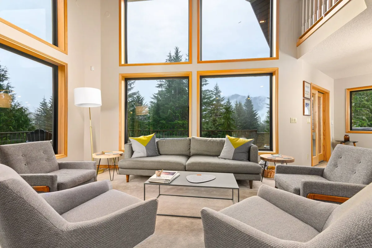 A spacious living room with large windows offering a stunning view of the mountains, complemented by a comfortable grey sofa, accent chairs, and a modern coffee table. The arrangement invites relaxation and conversation, while the expansive glass brings the outdoors in.