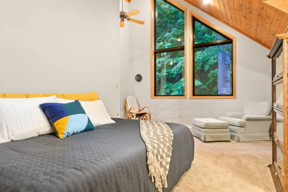 A serene bedroom scene with large windows framing views of greenery, a bed adorned with colorful pillows, and a comfortable armchair. The windows provide a clear visual connection to the surrounding natural environment, suggesting the ability to enjoy the changing seasons from the comfort of indoors.