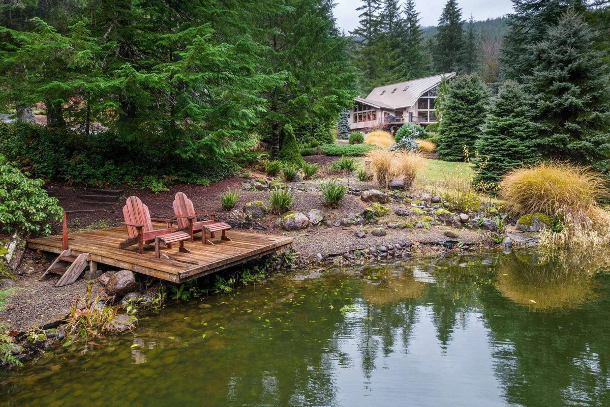 A broad wooden deck with chairs overlooks a tranquil pond surrounded by lush greenery, with the house visible in the background. The serene scene invites quiet contemplation or wildlife watching.