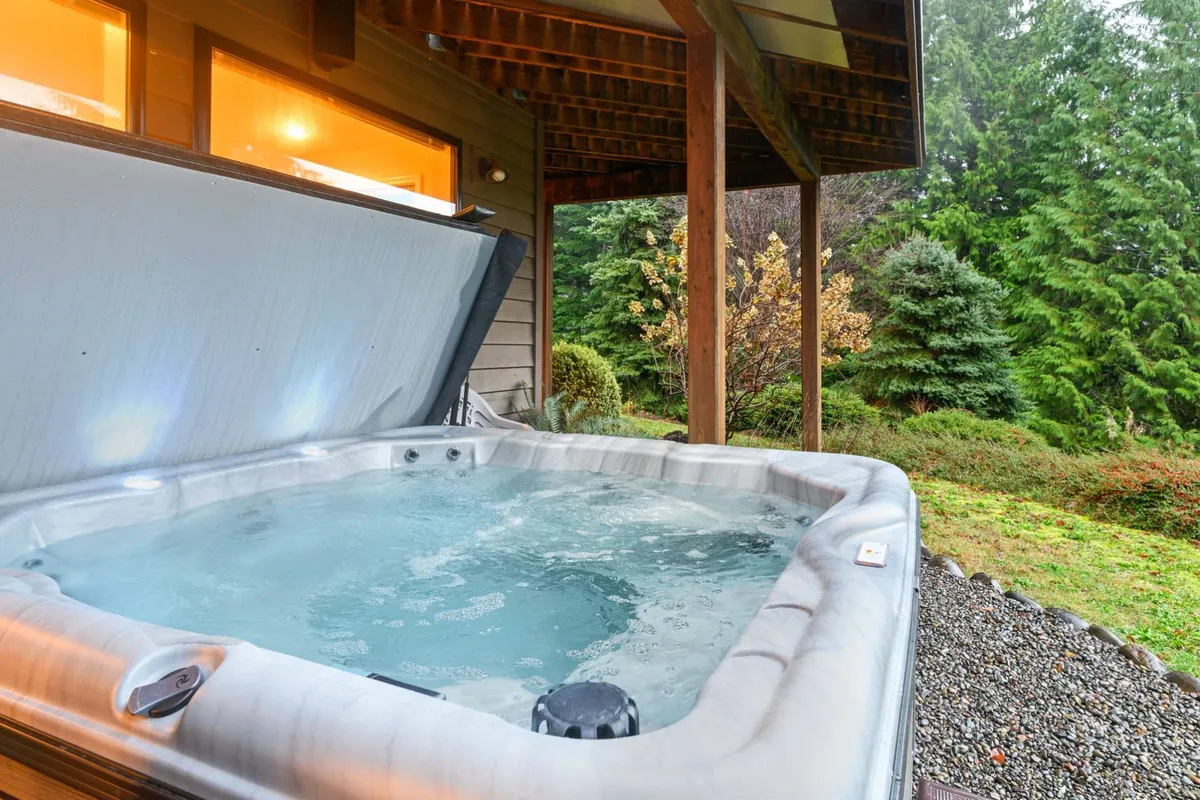 An outdoor hot tub nestled on a wooden patio, surrounded by lush greenery and partially shaded by a patio structure. The hot tub offers open views of the surrounding nature, evoking a sense of seclusion and relaxation in the mountain air.
