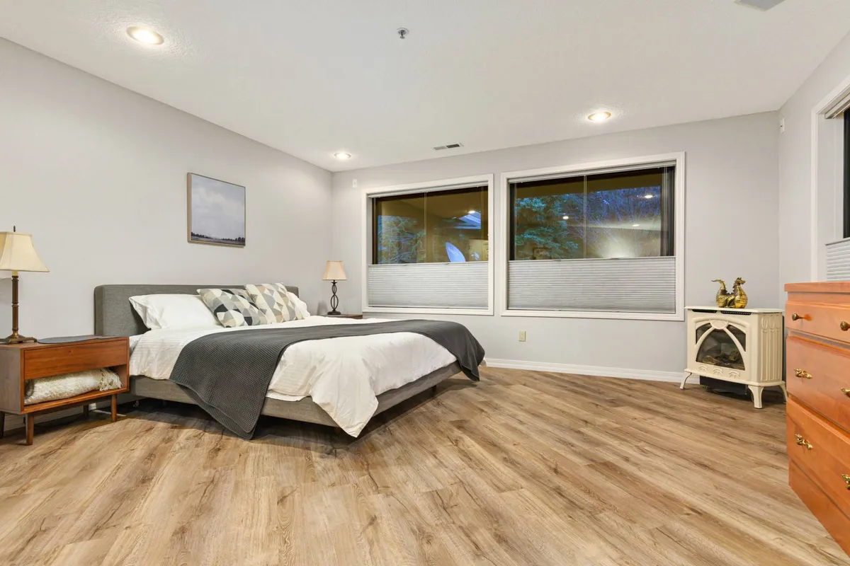 A cozy bedroom with large windows letting in streams of natural light, a bed with decorative pillows, a nightstand with a lamp, and a dresser. The image suggests the peacefulness of waking up to the sights and sounds of nature, with the vibrant outdoors always just a glance away.