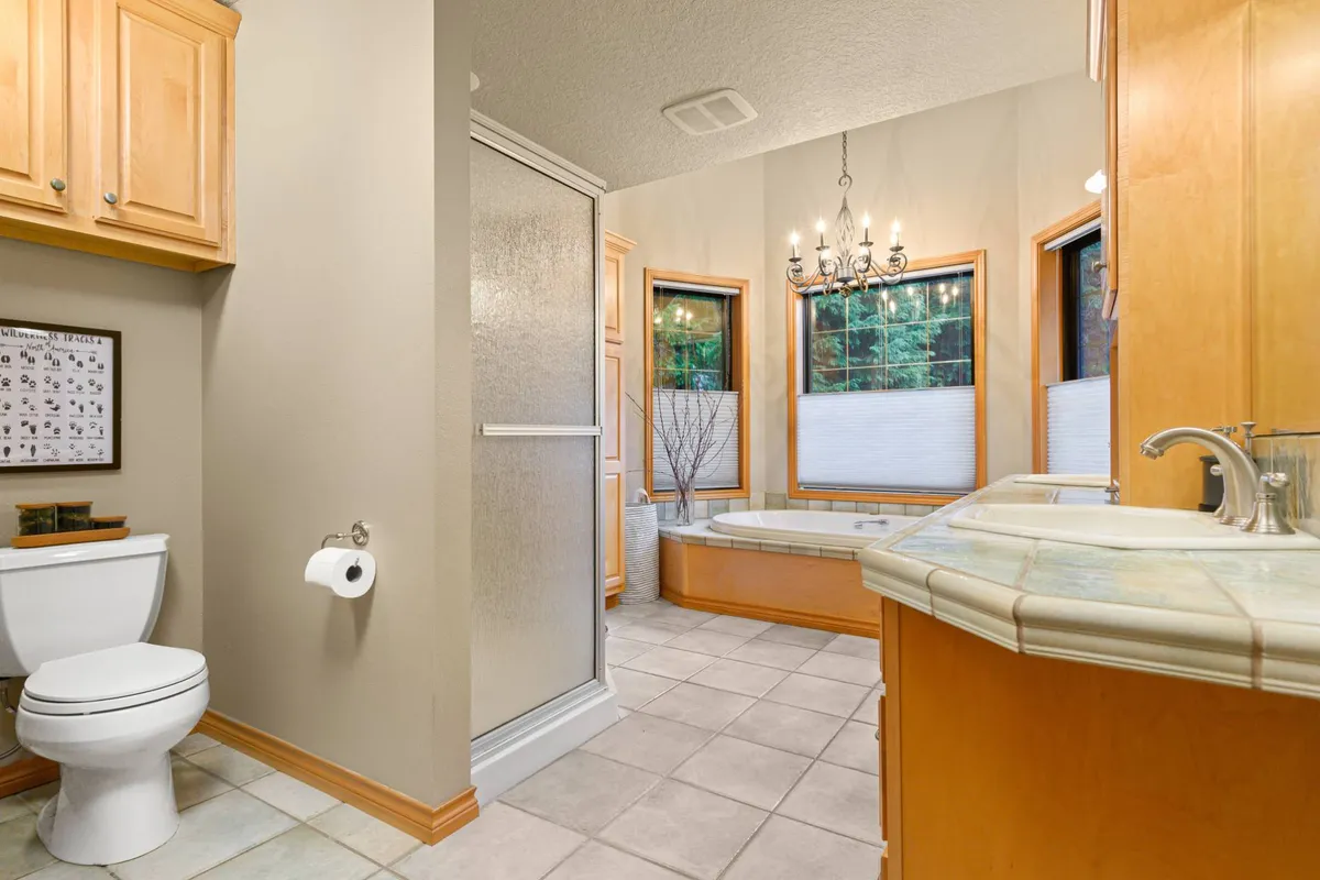 A bright, serene bathroom featuring a deep soaking tub, a glass-enclosed shower, a stylish chandelier, and a spacious vanity. The large windows flood the space with natural light, enhancing the sense of luxury and relaxation.