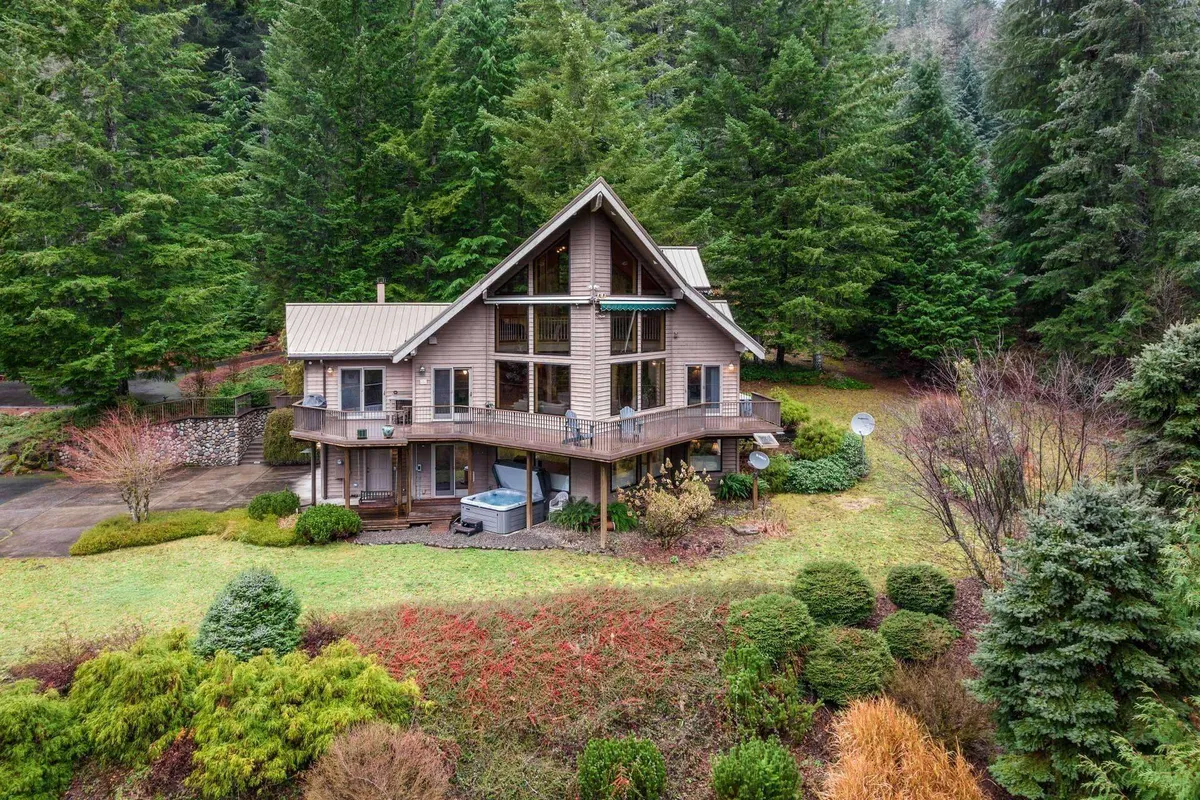 The image showcases the beautiful wooden exterior of a spacious house set amidst lush greenery, with large windows inviting in natural light. A generous deck extends from the house, perfect for relaxing or gathering with loved ones, and the tranquil surroundings hint at mountain views and ample outdoor space for activities.