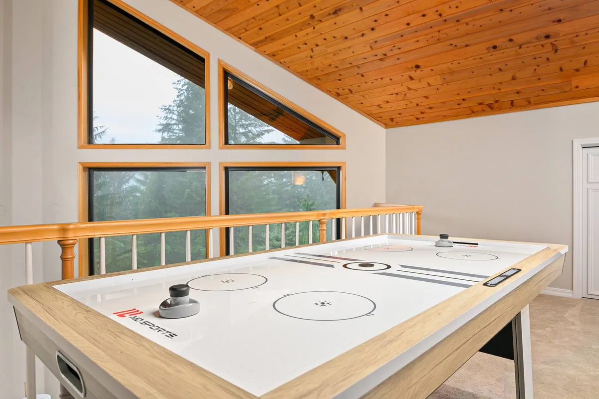 This image features a bright, inviting loft lounge with a prominent air hockey table near large windows. The room's wooden ceiling and ample natural light create an energetic, playful mood. There's comfortable seating visible in the background, suggesting space for both games and relaxation.